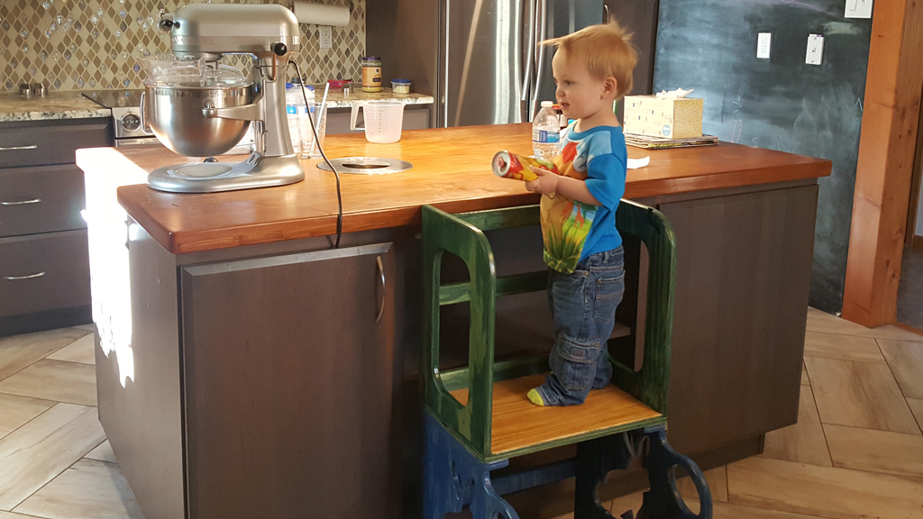 a happy toddler standing on a step stool in the kitchen helping to bake